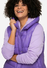 Gilet rembourré violet avec capuche, présentant une texture brillante et des boutons-pression. Porté sur un haut à manches longues violet clair avec des poignets côtelés.