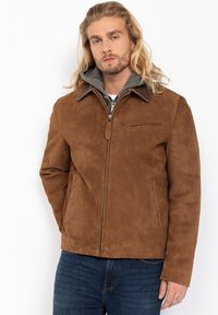 Uomo con lunghi capelli biondi che indossa una giacca di camoscio marrone sopra un hoodie grigio e jeans blu, in piedi con le mani ai fianchi.