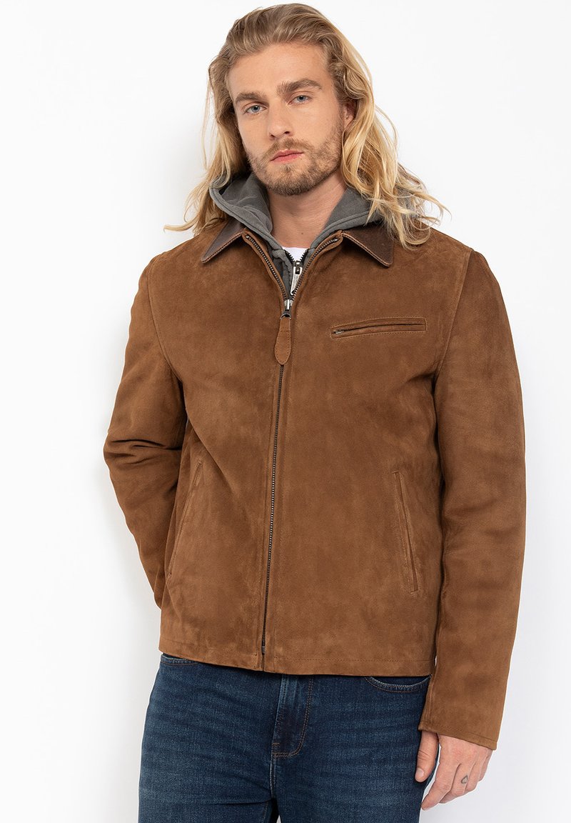 Man met lang blond haar, gekleed in een bruine suède jas over een grijze hoodie en blauwe spijkerbroek, staand met de handen naast zich.