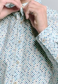 Hemd mit Knöpfen, das einen hellen Hintergrund mit einem geometrischen Muster in Blau, Türkis und Beige aufweist. Lange Ärmel mit geknöpften Manschetten.