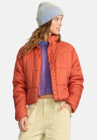 Gevoerde oranje jas met een cropped ontwerp. Heeft een hoge kraag en een elastische zoom. Gecombineerd met een blauw gestreept shirt en beige hoog-hoog taille broek.