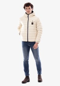 Giacca a vento di colore beige chiaro con cuciture orizzontali, chiusura con zip e cappuccio. Presenta una tasca sul petto con un'etichetta del marchio. Indossata con jeans blu.