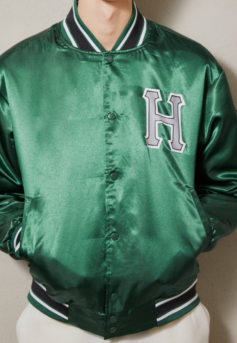 Grüne Satinschulterjacke mit weiß und schwarz gestreiftem Rippkragen und -bündchen. Hat ein großes graues "H" auf der linken Brust.