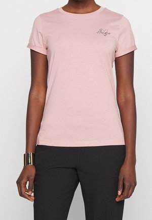 Kobieta ubrana w jasno-różową koszulkę z krótkim rękawem z podwiniętymi mankietami oraz czarne spodnie, ozdobiona szeroką złotą bransoletką na lewej ręce.