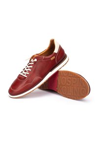 Zapatilla de cuero rojo con acentos blancos, puntera redonda, cordones y suela de goma marrón contrastante con marca en relieve.
