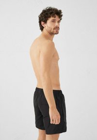 Shorts de bain noirs avec une taille élastique et une poche sur le côté, présentés sur un modèle masculin vu de côté, torse nu.