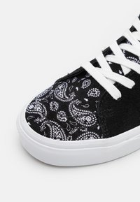 Černé tenisky s paisley vzorem v bílé barvě, semišový svršek, bílé tkaničky a gumová podrážka. Textura kontrastuje mezi látkou a semišem.