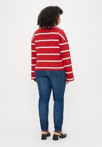 Haut long à rayures rouges et crème avec des manches larges, associé à un jean bleu ajusté et des chaussures à talons carrés noires. Tissu texturé.