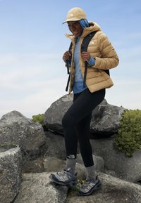 Chaqueta aislante beige con capucha, capa base azul, leggins negros y zapatos de senderismo grises sobre terreno rocoso. Correas de mochila visibles.