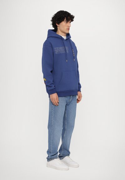 Felpa con cappuccio blu con stampa del logo, cappuccio con cordini e tasca frontale. Abbinata a jeans azzurri e sneakers bianche. Sfondo neutro.