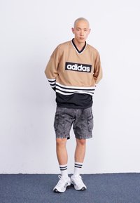 Μπέζ αθλητική μπλούζα Adidas με μαύρες λεπτομέρειες και ρίγες, συνδυασμένη με γκρίζες cargo βερμούδες. Μοντέλο που φοράει λευκά αθλητικά παπούτσια και αθλητικές κάλτσες.