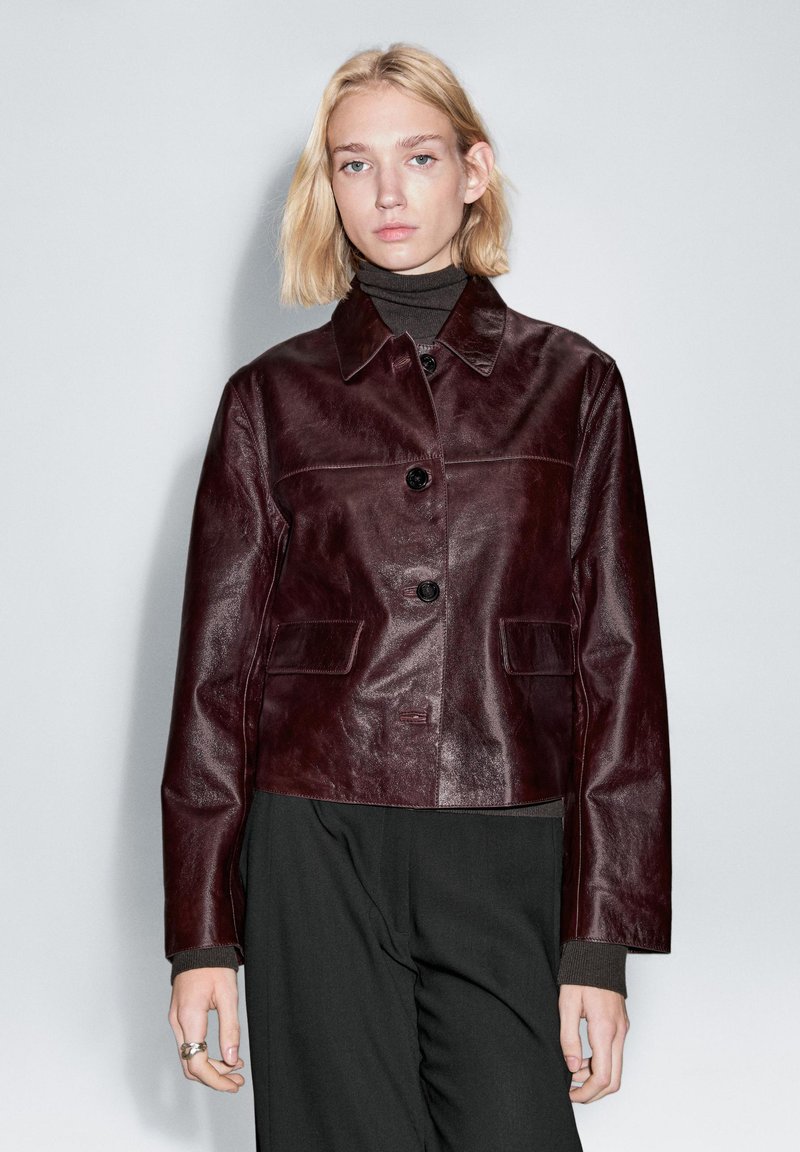 Bourgogne læderjakke med krave, lukning med knapper foran og to sidelommer. Glat tekstur med en let skinnende finish.