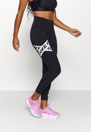 Kobieta w czarnych legginsach Nike Pro z wyróżniającym się białym logo i różowych butach do biegania Nike, stojąca na jednolitym białym tle.