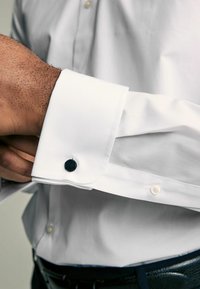 Weißes Hemd mit manschette und einem schwarzen sechseckigen Manschettenknopf. Glatte Stoffstruktur, mit Knopfverschluss und kontrastierenden Designelementen.