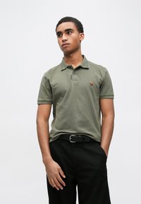 Olívovo zelené bavlnené polokošeľa s golierom, obsahujúca malý vyšívaný vzor. Nosené s čiernymi nohavicami a čiernym opaskom.