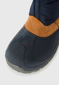 Gros plan sur une botte imperméable noire et marron avec un capuchon de protection texturé et une fermeture éclair sur le côté, sur fond blanc.