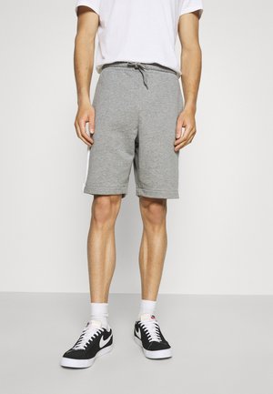 Mann trägt graue Shorts mit Kordelzug, weiße Kniestrümpfe, schwarze und weiße Nike-Sneaker und ein weißes T-Shirt, steht vor einem einfarbigen Hintergrund.