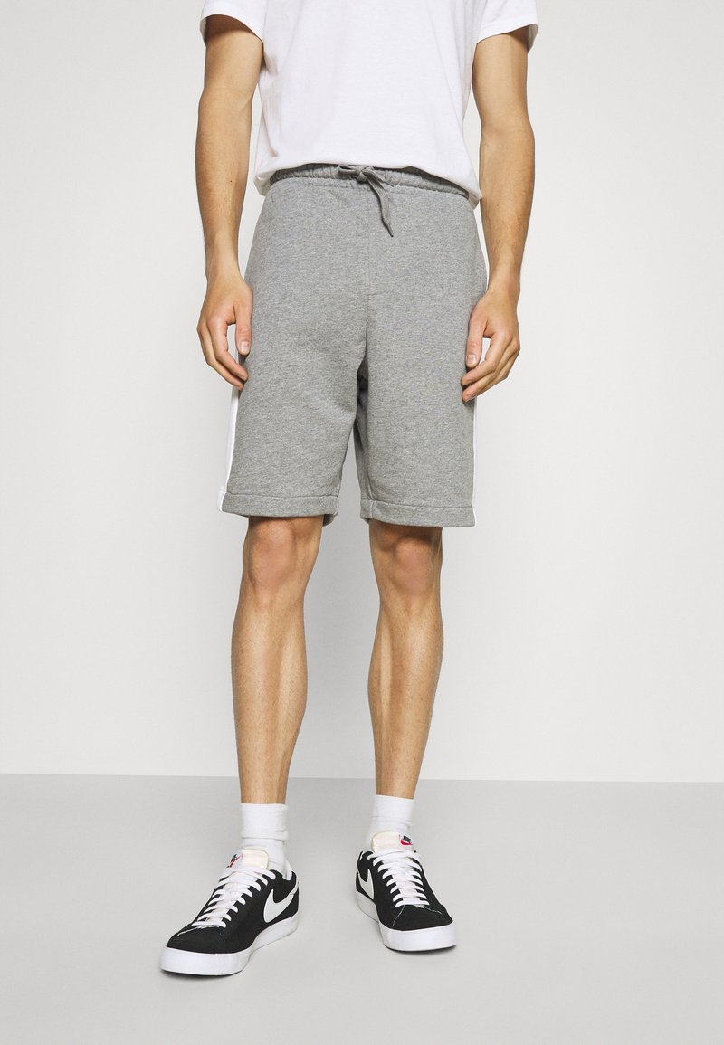 Homme portant un short gris à cordon, des chaussettes blanches montantes, des baskets Nike noires et blanches, et un t-shirt blanc, debout devant un fond uni.