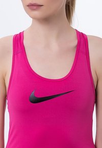 Fuksjowy top sportowy z okrągłym dekoltem, z czarnym logo Nike z przodu; gładka tekstura i model z wycięciem na plecach.