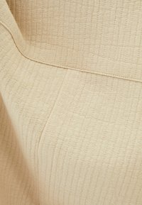 Beige ribbad tyg med vertikala sömmar och en söm som löper horisontellt över ytan, som visar textur och detalj.