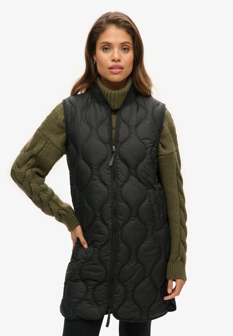 Schwarze gesteppe Weste mit hohem Kragen und Reißverschluss, über einem grünen Strickpullover mit Zopfmuster getragen.