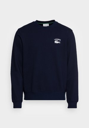 Sudadera azul marino con cuello redondo, puños y bajo acanalados. Presenta un logo de Lacoste bordado en blanco en el pecho. Textura de tejido suave.
