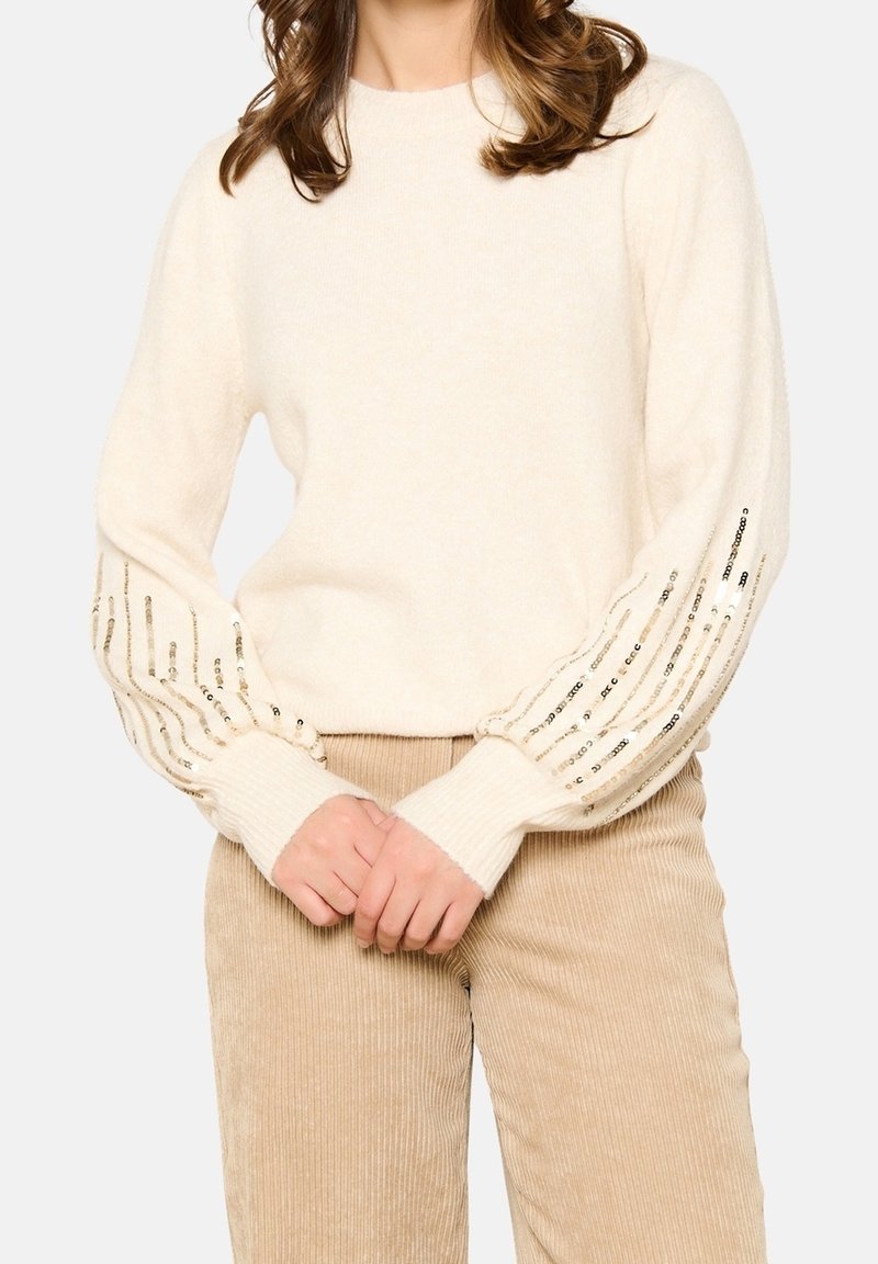 Femme portant un pull crème orné de rayures verticales en sequins brillants sur les manches et un pantalon en velours côtelé beige, les mains croisées devant elle.