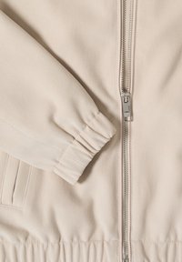 Veste beige à fermeture éclair avec un tissu texturé, des poignets élastiqués et une fermeture éclair métallique visible avec une tirette argentée.