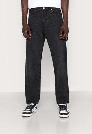 Man die zwarte rechte jeans en zwart-witte sneakers draagt, staande tegen een effen achtergrond.