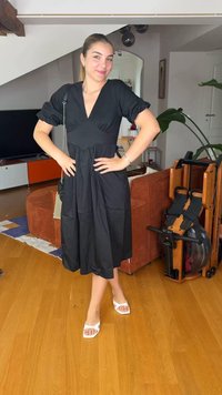 Γυναίκα με μαύρο μίντι φόρεμα και άσπρα σανδάλια στέκεται με τα χέρια στη μέση σε ένα σαλόνι με ξύλινο πάτωμα και κοντά βρίσκεται ένα όργανο κωπηλασίας.