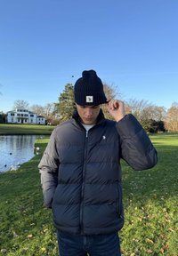 Noor mees kohendab tumerohelist mütsi, kandes tumedat sulejakki tiigi ääres, kus on pardid, roheline muru, puud ja taustal valge maja.