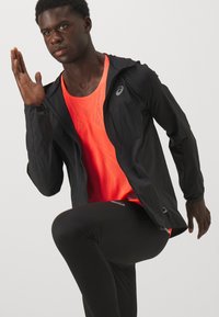 Mees mustas jooksujakus, heledalt oranžis spordisärgis ja mustades retuusides, tõstab paremat kätt ja tõstab vasaku põlve jooksuasendis.