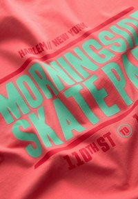 Koralfarvet t-shirt med fed mintgrøn tekst, der står "MORNINGSIDE SKATEPARK" med "HARLEM// NEW YORK" og "110TH ST TIL" detaljer. Blødt bomuldsmateriale.