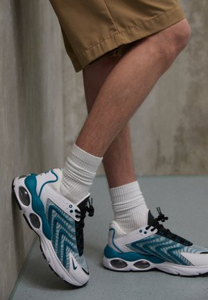 Persona con pantaloncini color beige, calzini bianchi e sneakers color verde acqua e bianchi appoggia un piede contro una parete grigia in un ambiente interno minimalista.