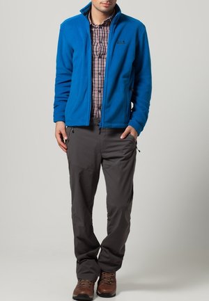 Mann trägt eine blaue Fleecejacke über einem rot-weiß karierten Hemd, graue Hose und braune Wanderschuhe, steht vor einfarbigem Hintergrund.