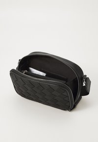Zwarte crossbody tas met een geweven voorpaneel, open ritssluiting die de stoffen binnenkant laat zien en een gedeeltelijk zichtbare witte papier binnenin.