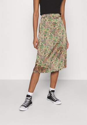 Personne portant une jupe midi fleurie colorée, un haut noir sans manches, des chaussettes blanches et des baskets montantes noires, debout devant un fond uni.