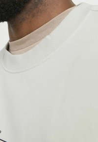 Lichtcrème sweatshirt met een geribde ronde halslijn, gecombineerd met een beige onderhemd. Zachte stoftextuur en minimalistische designelementen.