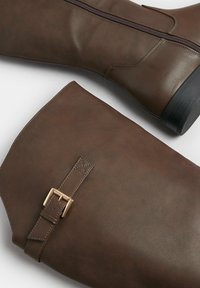 Stivali marroni in pelle al ginocchio con cerniera laterale e un accento in fibbia sul gambale. Texture liscia con finitura opaca. Suola in gomma nera.