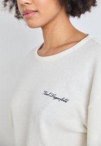 Sweatshirt branco texturizado com gola redonda e um logótipo bordado preto no peito. O tecido tem um padrão tecido.