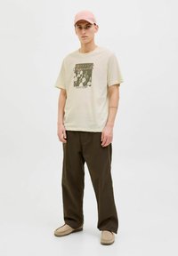 Beige tričko s grafikou kaktusu, v kombinaci s tmavě zelenými kalhotami. Model nosí růžovou čepici a světle hnědé slip-on boty. Jednoduchý a neformální design.