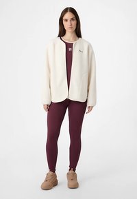 Lichtcrèmekleurig fleecejack met ritssluiting over een bordeauxrood bodysuit en legging, gecombineerd met beige sneakers. Opvallend logodetail op het jack.