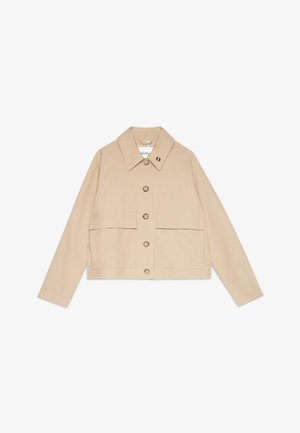 Giovane donna con capelli corti e ricci che indossa un cappotto beige, un vestito crema con una piccola stampa di frutta rossa e una sciarpa bianca con pois neri e motivo floreale.