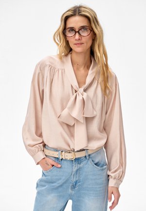 Kobieta z długimi blond włosami, nosząca okulary w oprawkach w kolorze żółwia, bladoróżową bluzkę z kołnierzykiem wiązanym na kokardę, jasnoniebieskie dżinsy oraz beżowy pasek.