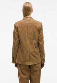 Blazer en velours côtelé marron avec des côtes verticales ; présente un col cranté et des manches à poignets boutonnés. Présenté de dos sur un fond blanc.