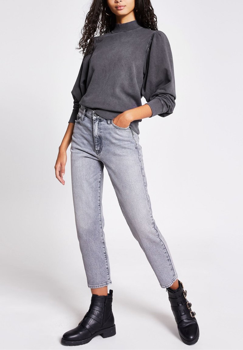 Grauer Sweatshirt mit hohem Kragen und Puffärmeln, kombiniert mit hoch taillierten, hellgrauen Jeans. Schwarze Knöchelstiefel mit Riemenakzenten.