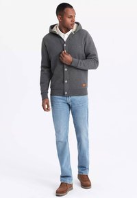 Grå cardigan med hætte, knapper foran, lys foring, båret over en hvid skjorte, kombineret med blå jeans og brune støvler.
