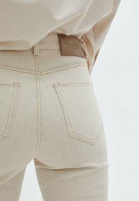 Jean en denim beige taille haute, avec poches arrière, une étiquette en cuir à la taille et des détails de couture subtils.