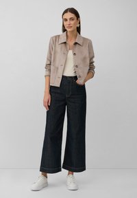 Giacca in suede beige con ampio collo e bottoni frontali, abbinata a pantaloni wide-leg in denim scuro e sneakers bianche.
