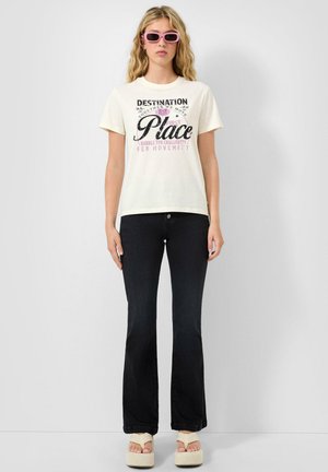 Jeune femme aux longs cheveux blonds portant des lunettes de soleil roses, un t-shirt graphique blanc, un jean évasé noir et des sandales compensées beiges.